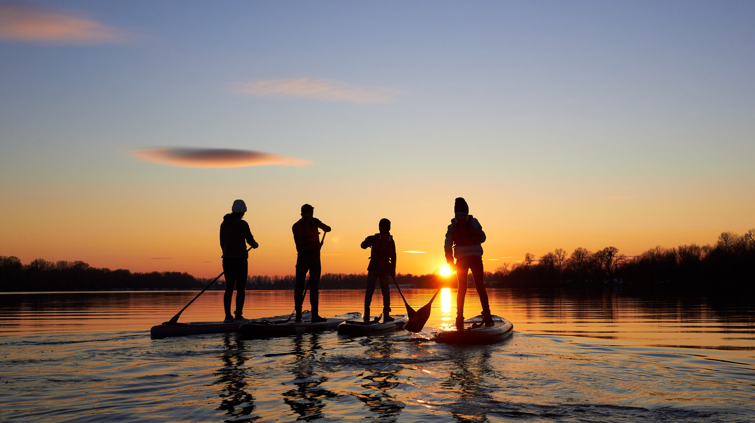 Silhouettes of the people on stand up paddle board at dusk on a flat quiet winter river with beautiful sunset colors